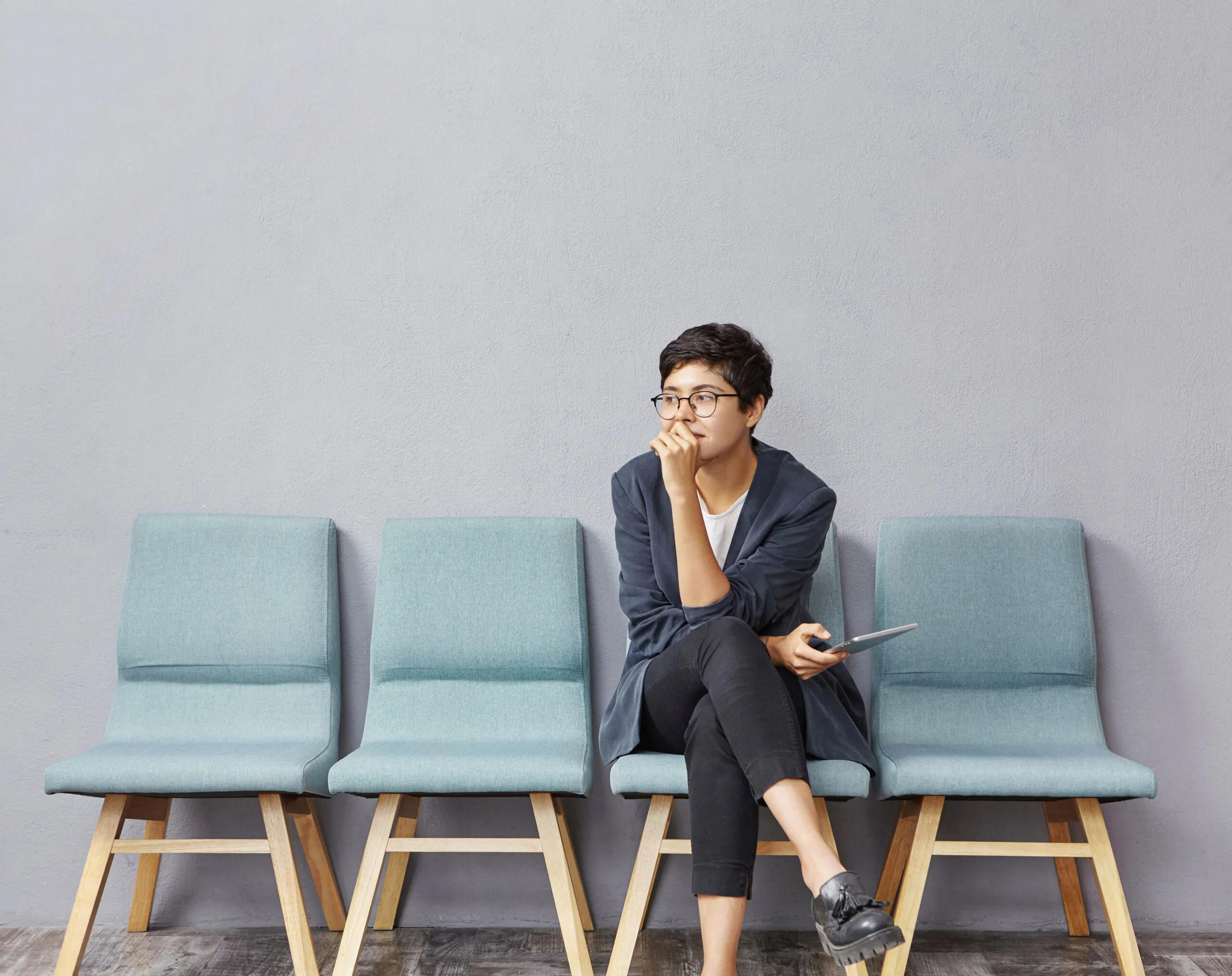 Picture of attractive fashionable young female designer wearing trendy clothes and eyeglasses sitting on chair in hall, keeping legs crossed, feeling nervous before job interview, biting her nails Proceso de reclutamiento de personal
