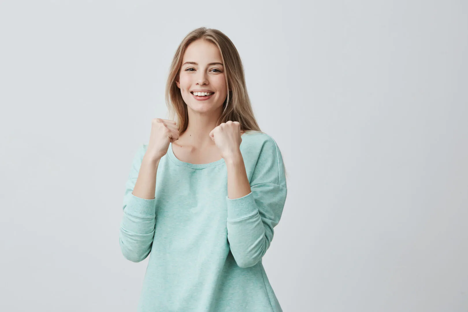 Lucky young female employee rejoicing success at work, smiling broadly, keeping fists clenched. Beautiful blonde girl in light blue sweater feeling happy and excited posing against gray background Retos para Recursos Humanos en el 2023